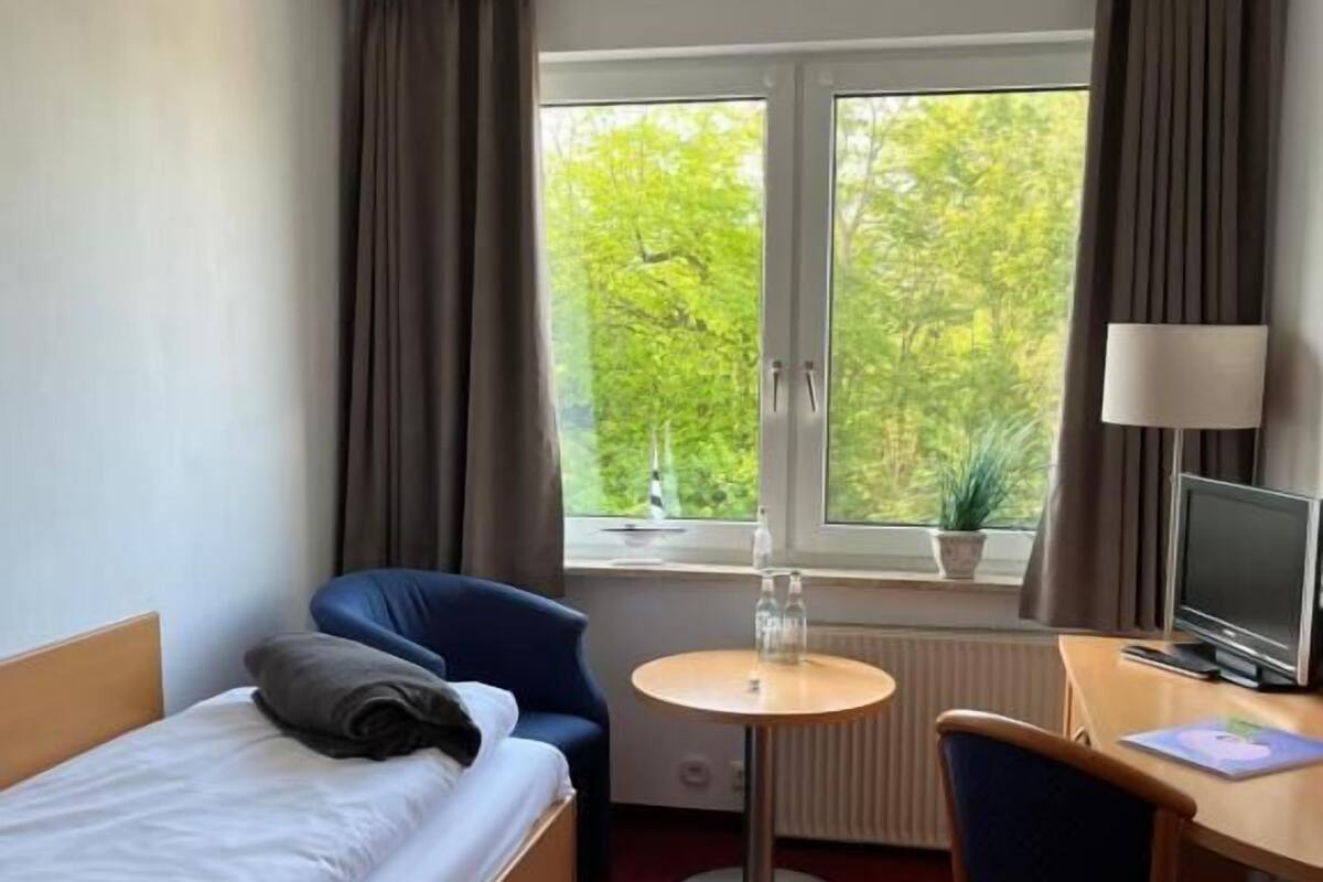 Ein Hotelzimmer mit einem Bett, einem Schreibtisch, einem Stuhl, einem kleinen Tisch, einer Lampe und einem Fenster mit Vorhängen.