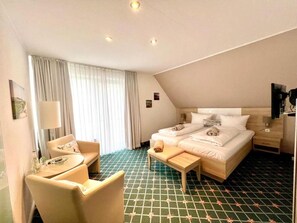 Una habitación de hotel con cama, una silla, una mesita, una lámpara y una ventana con cortinas.
