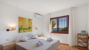 A bedroom with a bed, bedside tables, a wall-mounted air conditioner, a window with a view, and a painting on the wall. - Fantastic Villa with Pool and Spectacular Sea Views (Sa Riera)