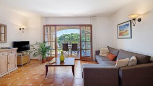 A living room with a sofa, TV, and a table with a vase. There is a balcony with chairs and a view of greenery. - Fantastic Villa with Pool and Spectacular Sea Views (Sa Riera)
