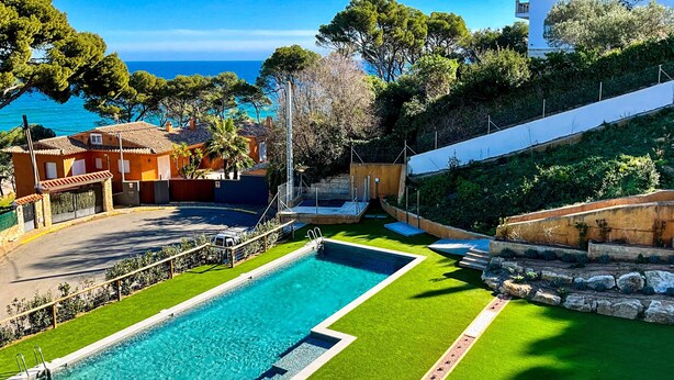 Ein Swimmingpool mit klarem, blauem Wasser, umgeben von grünem Kunstgrün, einer Steinmauer und Blick aufs Meer.