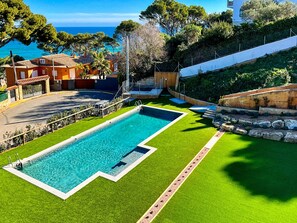Una piscina con agua azul cristalina, rodeada de césped artificial verde, un muro de piedra y vistas al mar.