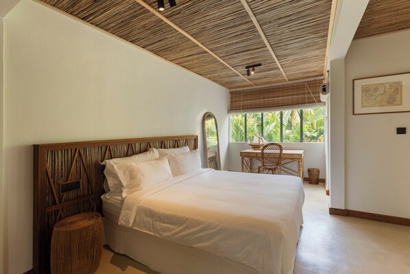 A bedroom with a large bed, a wooden headboard, a small table, and a window with a view of greenery.