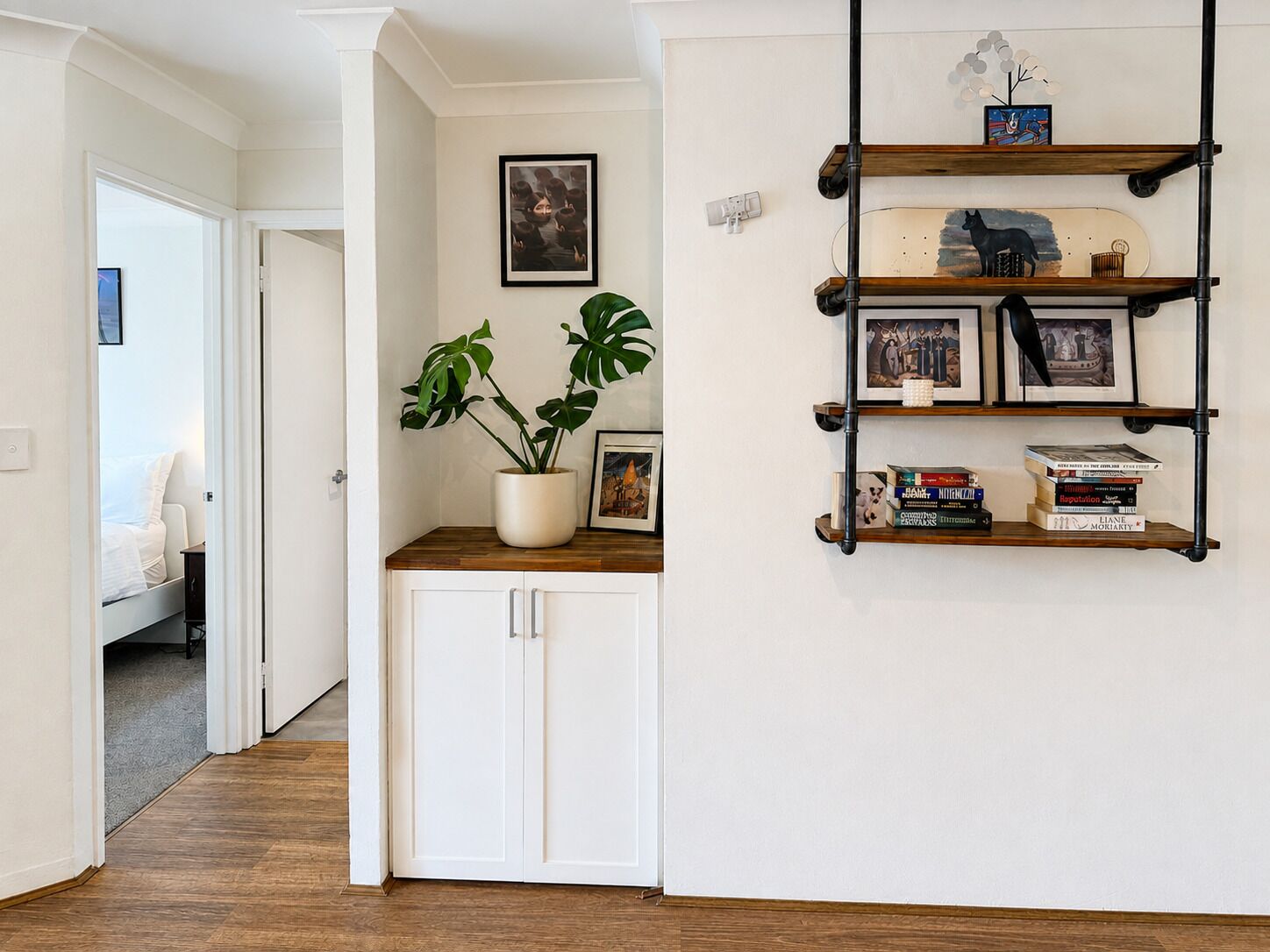 A hallway with a white door, a potted plant, and a wall-mounted shelf with books and framed pictures.