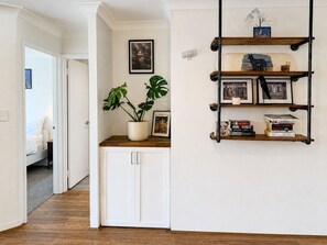 A hallway with a white door, a potted plant, and a wall-mounted shelf with books and framed pictures.