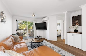 A modern living room with a leather sofa, a coffee table, a flat-screen TV, and a view of the outdoors through large windows. - Welcome to Magical McAtee, a stylish resort-style apartment just 5 minutes from the heart of Fremantle. <br>Relax by the sparkling pool, enjoy a luxurious king bed, free parking, fast Wi-Fi and a fully equipped kitchen. <br>Perfect for couples, famil (Fremantle)