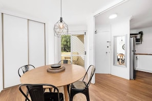 A dining area with a round table, chairs, a hanging light fixture, and a view through a window to an outdoor space. - Welcome to Magical McAtee, a stylish resort-style apartment just 5 minutes from the heart of Fremantle. <br>Relax by the sparkling pool, enjoy a luxurious king bed, free parking, fast Wi-Fi and a fully equipped kitchen. <br>Perfect for couples, famil (Fremantle)