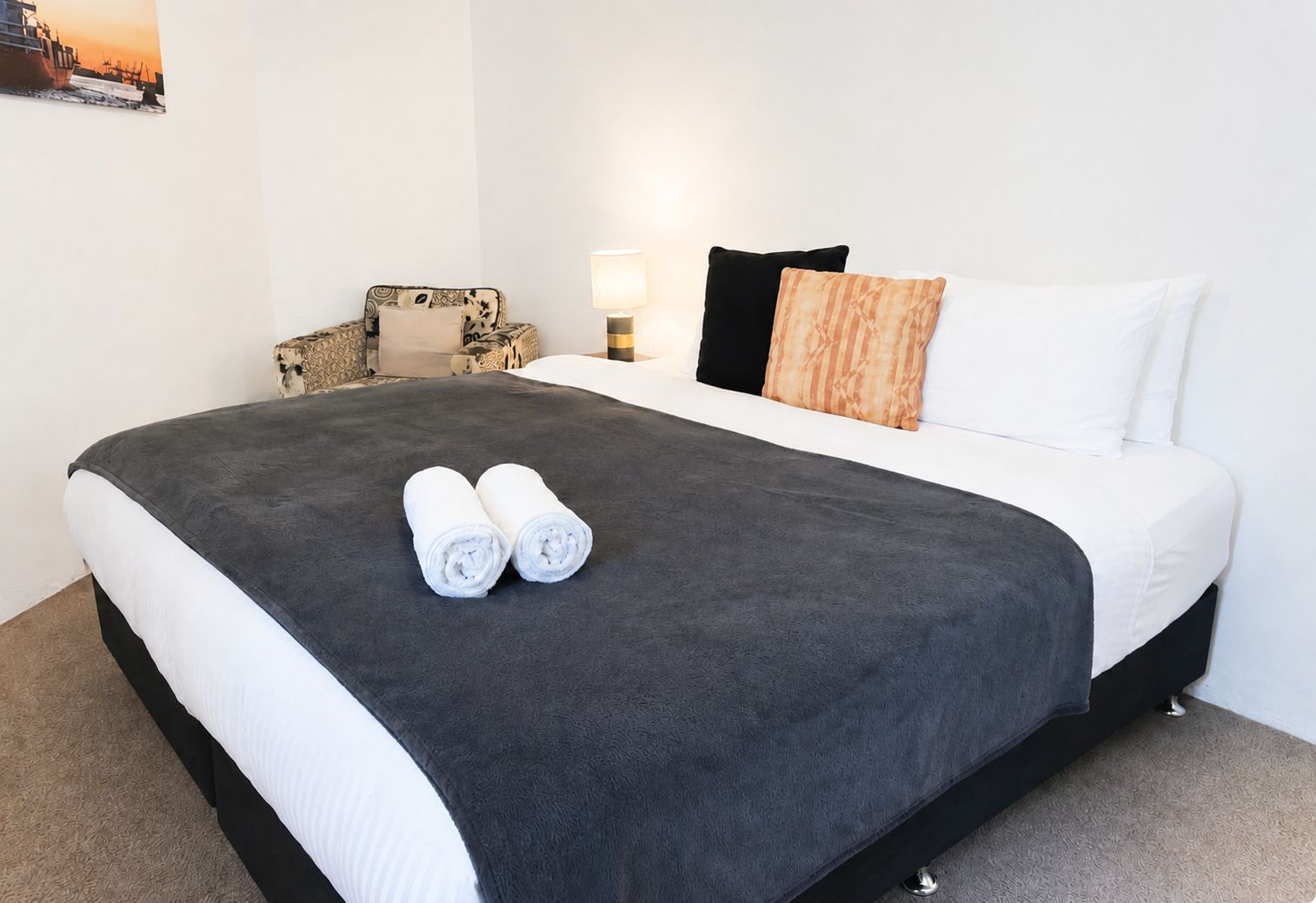 A hotel room with a bed, pillows, a chair, a lamp, and a painting on the wall.