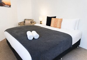 A hotel room with a bed, pillows, a chair, a lamp, and a painting on the wall. - Stylish two-bedroom, resort-style apartment for up to four guests, five minutes from Fremantle city centre. Unwind with a king bed, two private balconies, shared pool, fast Wi-Fi, split-system air-conditioning and heating, and free undercover parking (Fremantle)