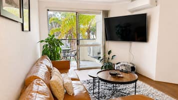 A living room with a leather sofa, a coffee table, a TV, and a balcony with plants.
