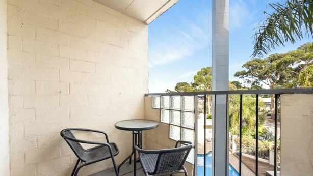 Un balcón con dos sillas y una mesita, vista a una piscina y un muro de ladrillo.