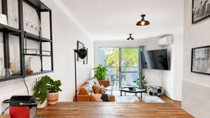 Una sala moderna con suelo de madera, un sofá, una mesa de centro, un televisor montado en la pared y un balcón con plantas.