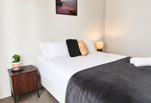 A neatly made bed with white bedding, a dark grey blanket, and multiple pillows. There is a small bedside table with a lamp and a potted plant. - Stylish two-bedroom, resort-style apartment for up to four guests, five minutes from Fremantle city centre. Unwind with a king bed, two private balconies, shared pool, fast Wi-Fi, split-system air-conditioning and heating, and free undercover parking (Fremantle)