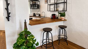 Cocina con mostrador de madera, dos taburetes de bar, una estantería metálica negra con cristalería y una planta en maceta.