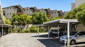 Un área de estacionamiento con marquesinas cubiertas, un coche blanco y edificios de apartamentos al fondo.