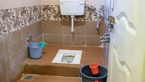 A small bathroom with a toilet, a bucket, a blue stool, and a shower area. - hari krupa bhakta niwas (Jejuri)