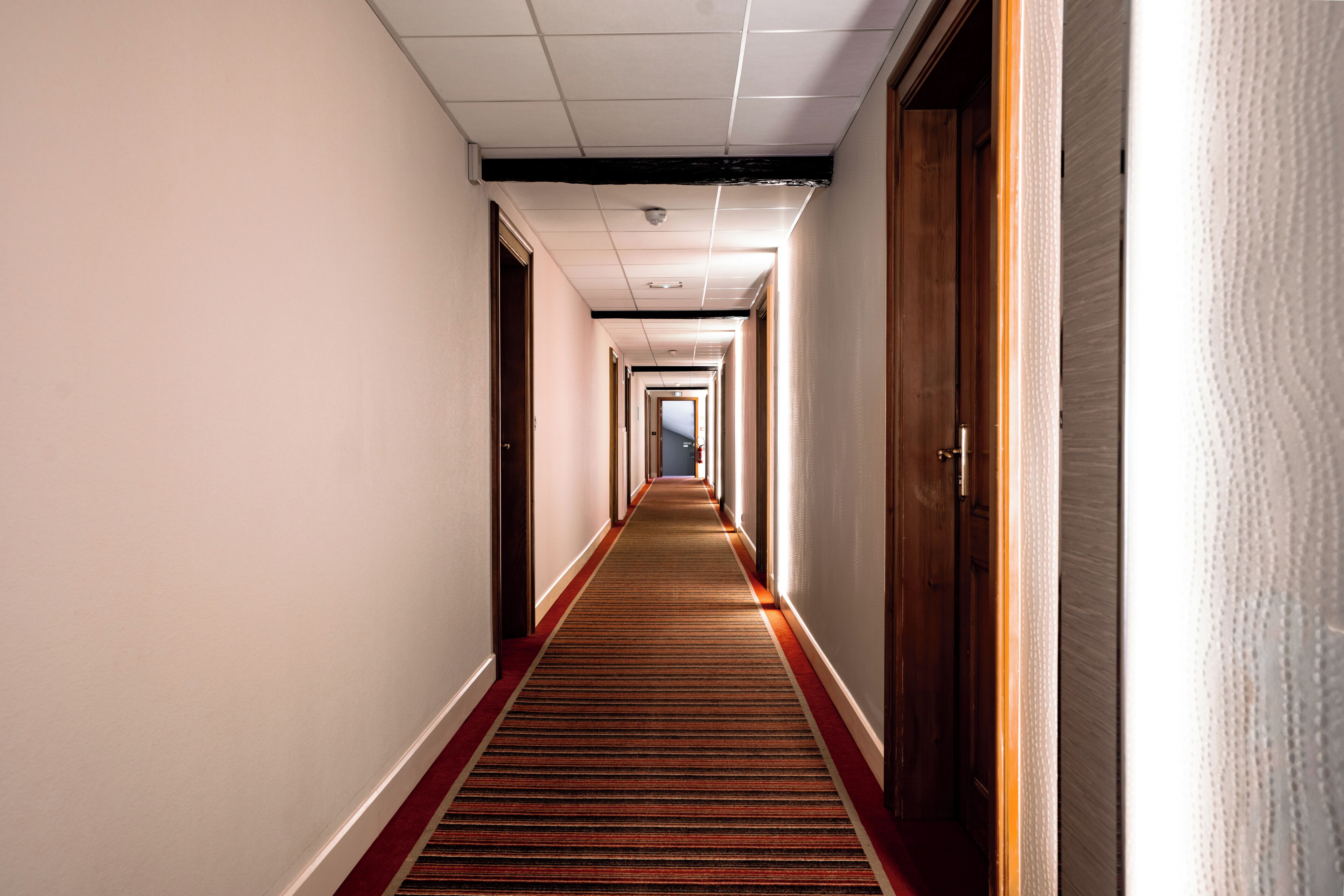 A hotel corridor with a carpeted floor, wooden doors, and a ceiling with recessed lighting.