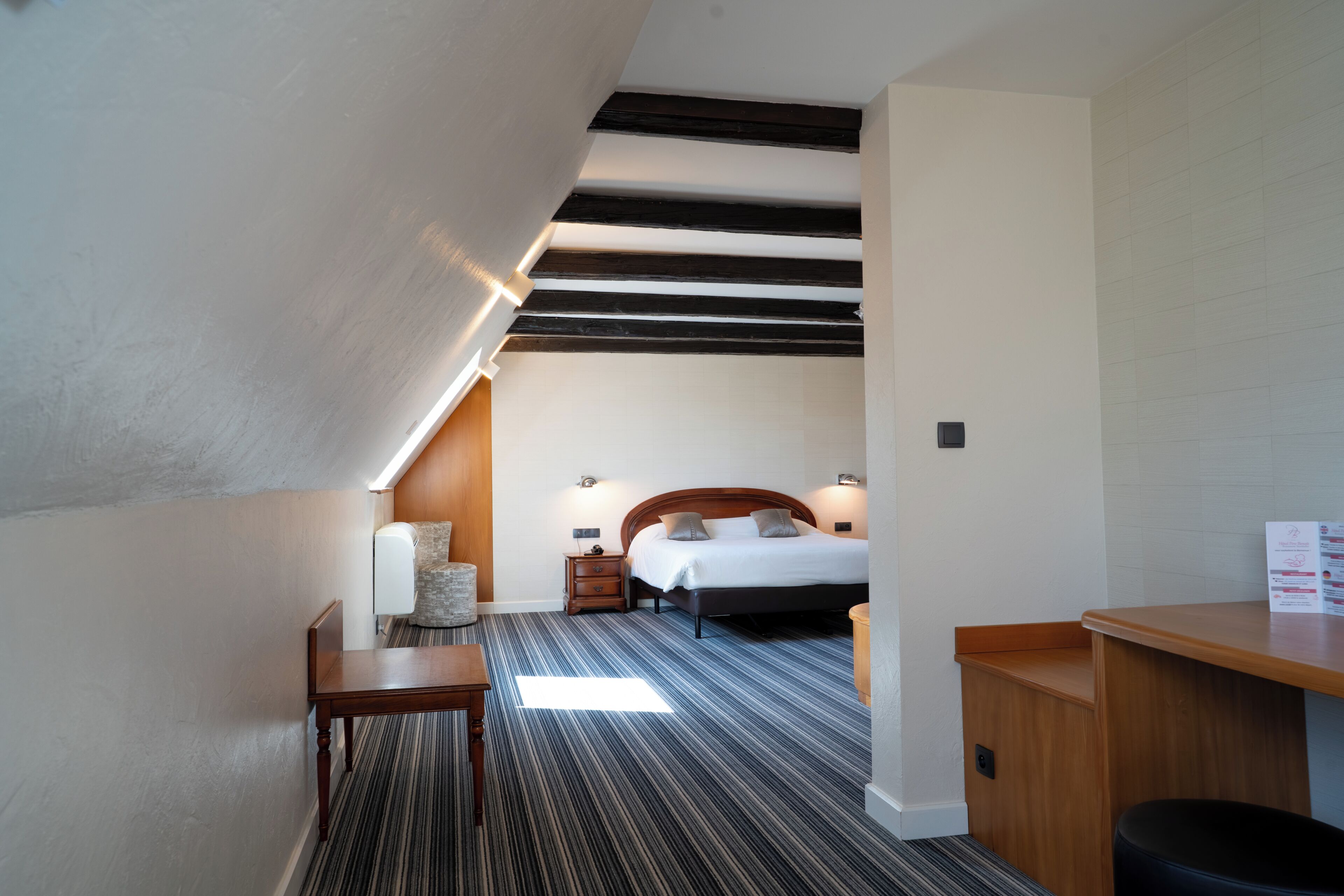 A hotel room with a sloped ceiling, a bed with pillows, a bedside table, a nightstand, and a small seating area with a chair.