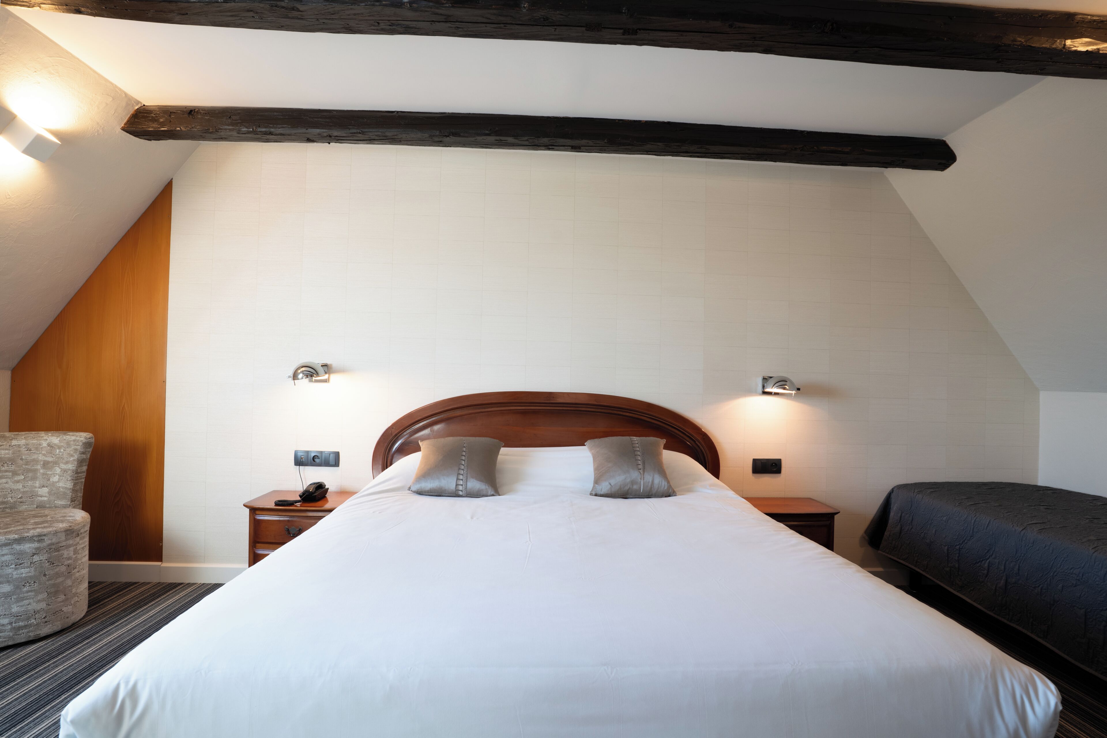 Una habitación de hotel con cabecera de madera, una cama individual con ropa blanca, una mesita de noche con teléfono y una silla.