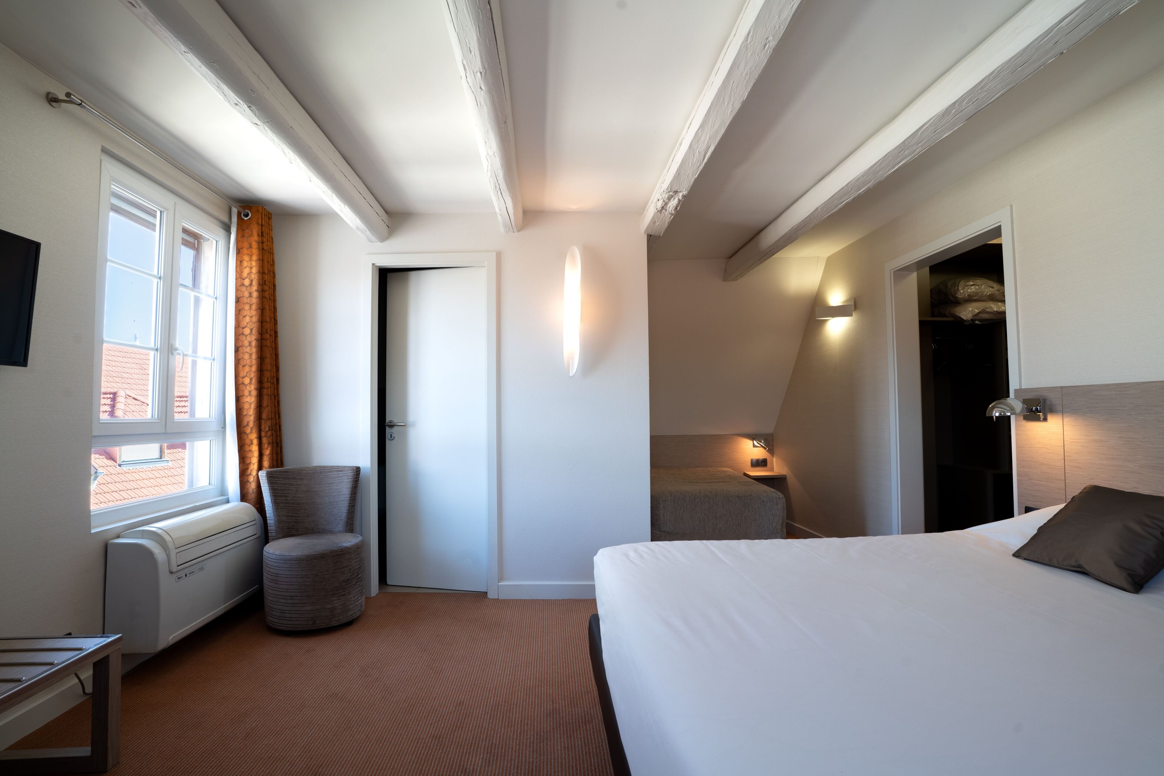 Ein Hotelzimmer mit Bett, Sessel, Schreibtisch, Heizkörper, einem Fenster mit Vorhängen, einer Tür und einem Schrank.