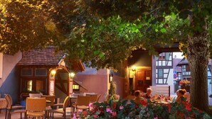 An outdoor seating area with tables and chairs under a tree, surrounded by flowering plants. - Hôtel Restaurant Père Benoît (Entzheim)