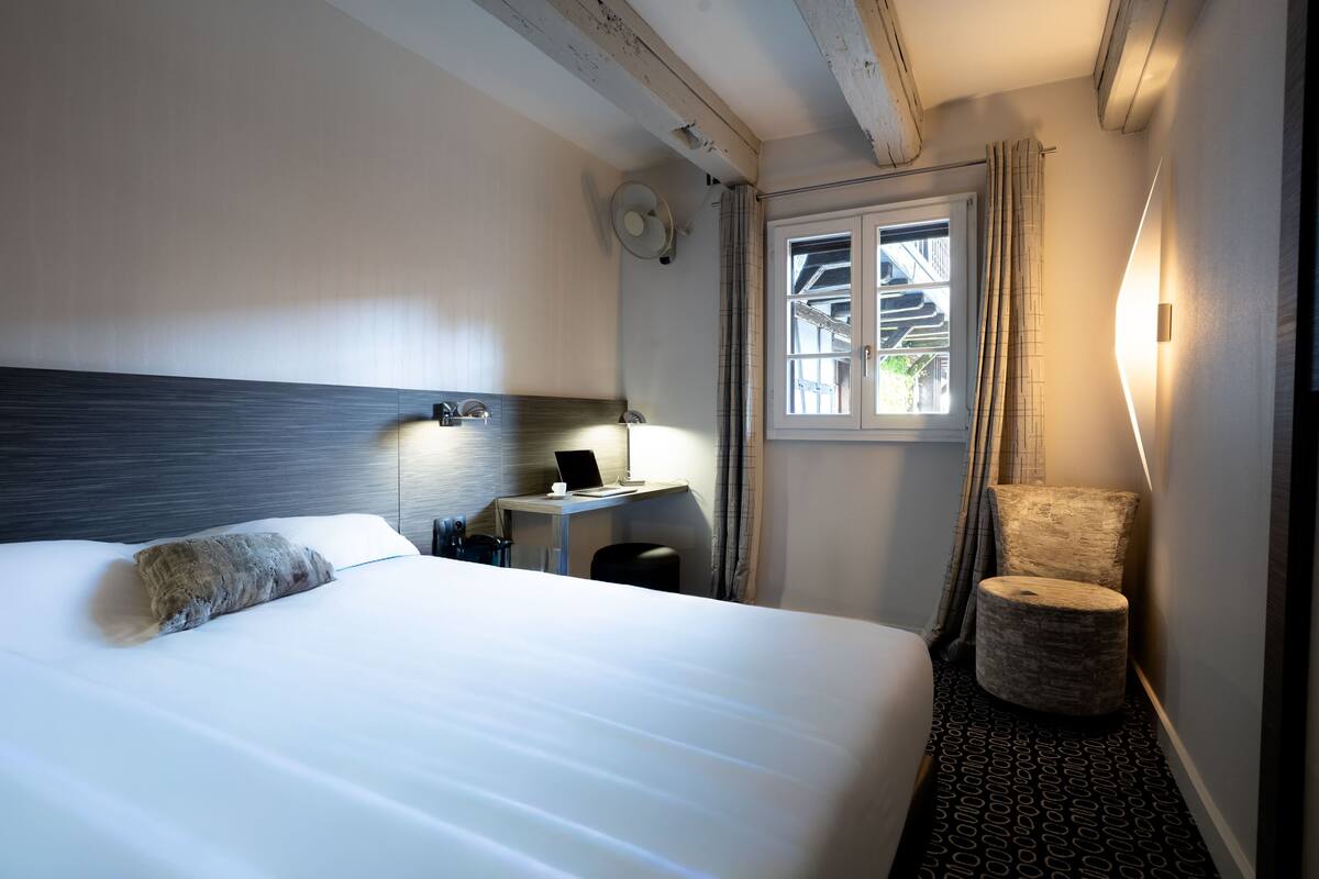 Habitación de hotel con cama, escritorio con computadora portátil, silla, ventana con cortinas y luz en la pared.