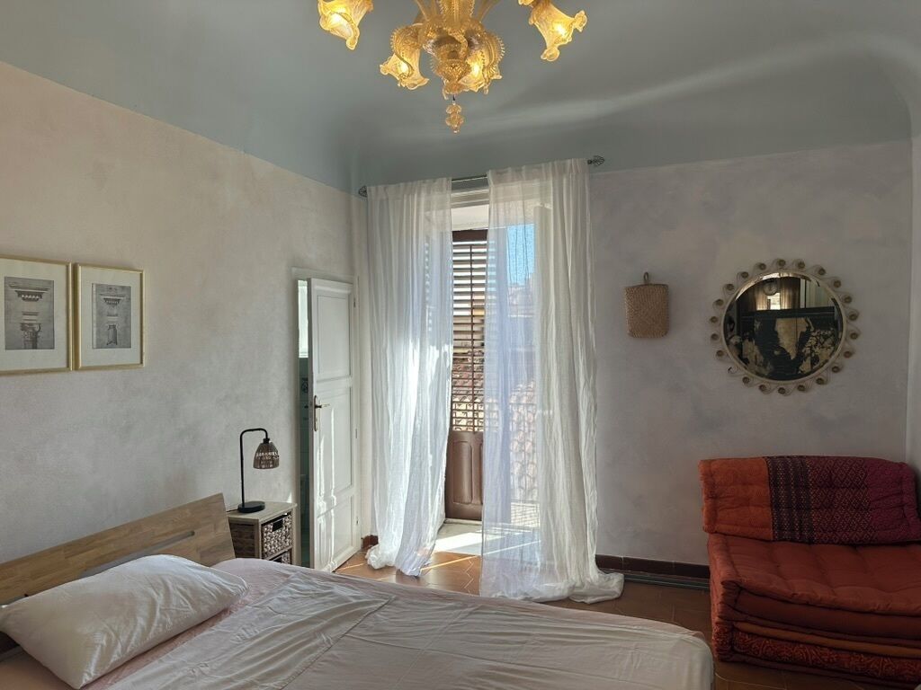 Una camera da letto con un letto, una lampada da comodino, una finestra con tende leggere, uno specchio rotondo e una sedia rossa.