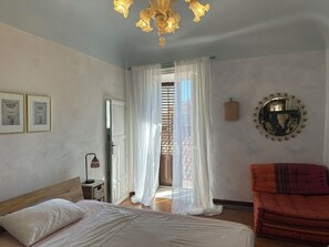 A bedroom with a bed, a bedside lamp, a window with sheer curtains, a round mirror, and a red chair.