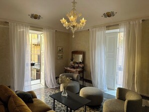 A living room with a sofa, ottomans, a table with a vase, a chandelier, and a desk with books and a mirror.