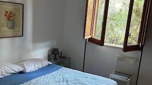 Un dormitorio con cama, una silla, una mesita de noche, una lámpara, una ventana con contraventanas abiertas y un cuadro en la pared.