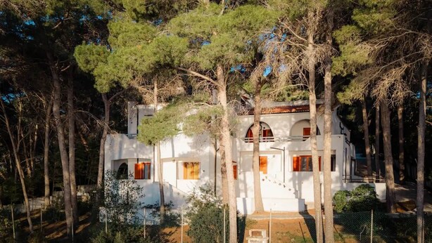 Una villa blanca con una escalera que lleva a un balcón, rodeada de árboles altos.