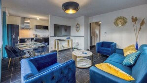 Un salon moderne avec un coin repas, une cuisine à l’arrière-plan et un canapé bleu agrémenté de touches dorées.