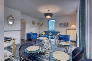 A dining area with a glass table set for six, a blue sofa in the background, and a chandelier above. - Le Donjon Bleu | Quiet / Mountain view (Varces-Allières-et-Risset)