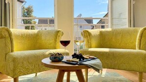 Un balcón con dos sillas amarillas tapizadas, una mesita redonda con un recipiente de bocadillos, dos copas de vino y vistas a edificios a través de amplias ventanas.