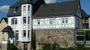 Una casa tradicional de entramado madera con base de piedra y sección superior blanca, destacando una torre prominente y un muro de piedra con una puerta.