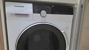 Eine Waschmaschine von Thomson mit Frontlader und Bedienfeld, in einem Raum mit Fliesenboden und einem gewebten Korb darauf.