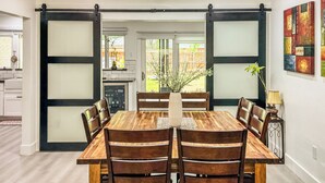 Een eetgedeelte met een houten tafel en stoelen, een vaas met planten, en een schuifdeur naar een keuken.