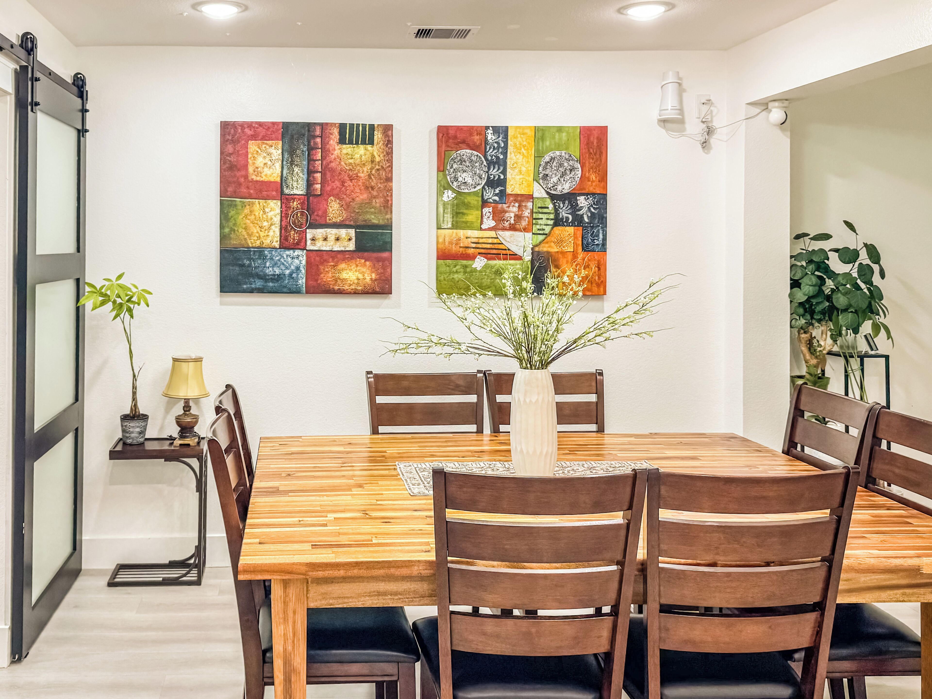 A dining area with a wooden table, six chairs, two abstract paintings, a vase with flowers, a lamp, and a potted plant.
