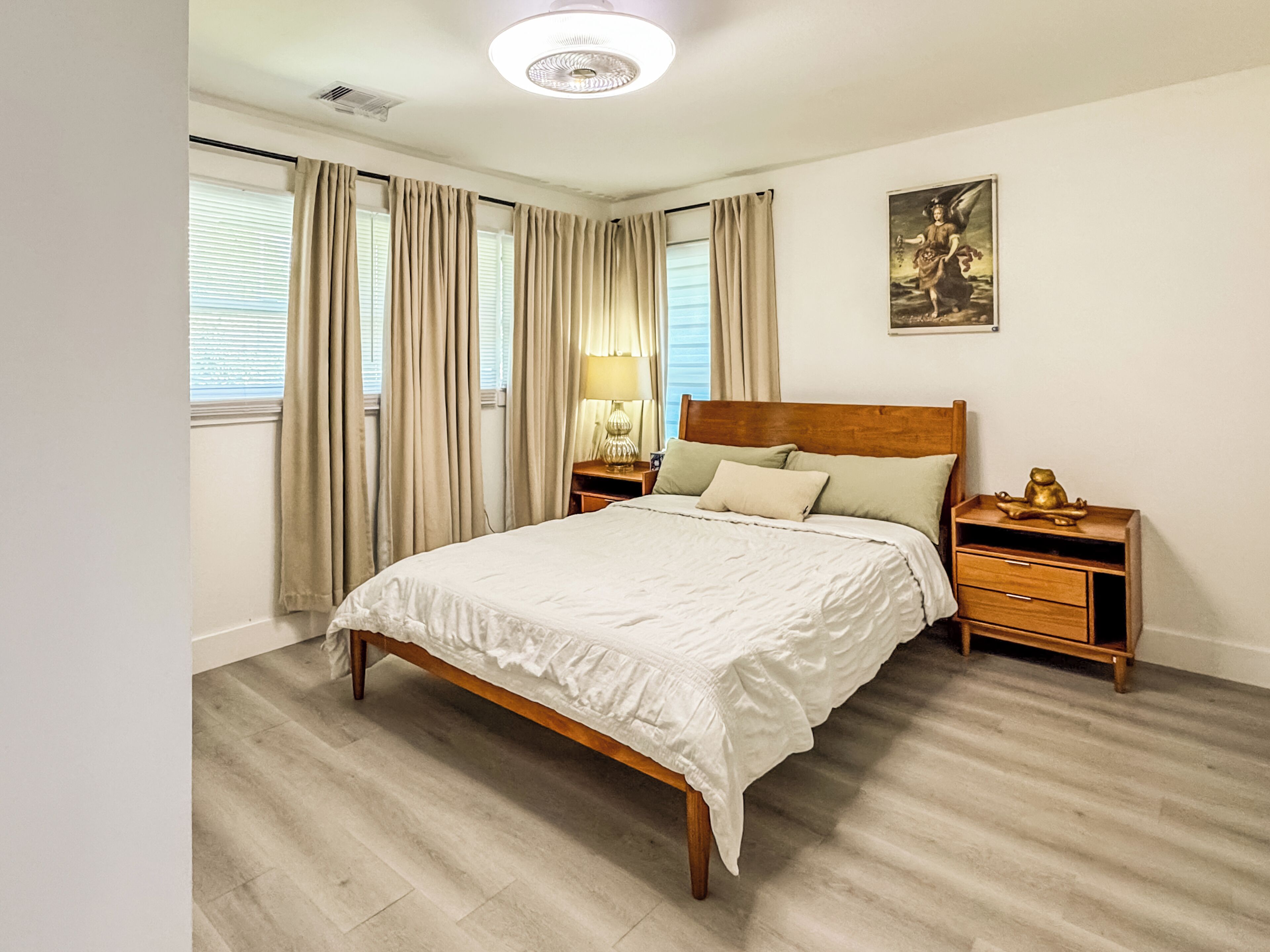 Quarto com cama de madeira, cortinas bege, mesa de cabeceira com abajur e quadro na parede.