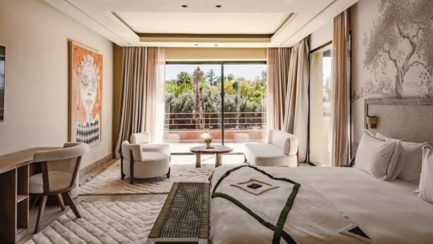 Ein Hotelzimmer mit einem großen Bett, zwei Sesseln, einem Schreibtisch und Blick auf Bäume durch ein großes Fenster.
