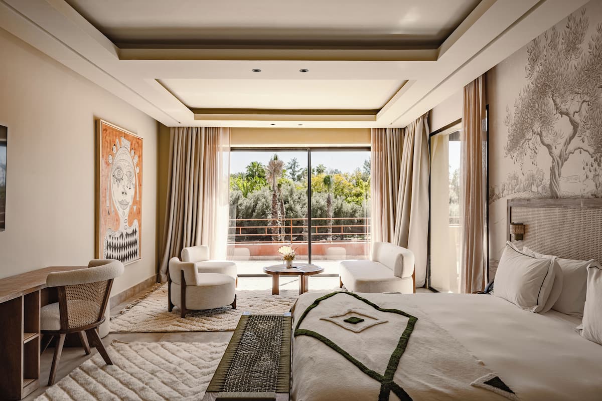 Une chambre d’hôtel avec un grand lit, deux fauteuils, un bureau et une vue sur les arbres par une grande fenêtre.