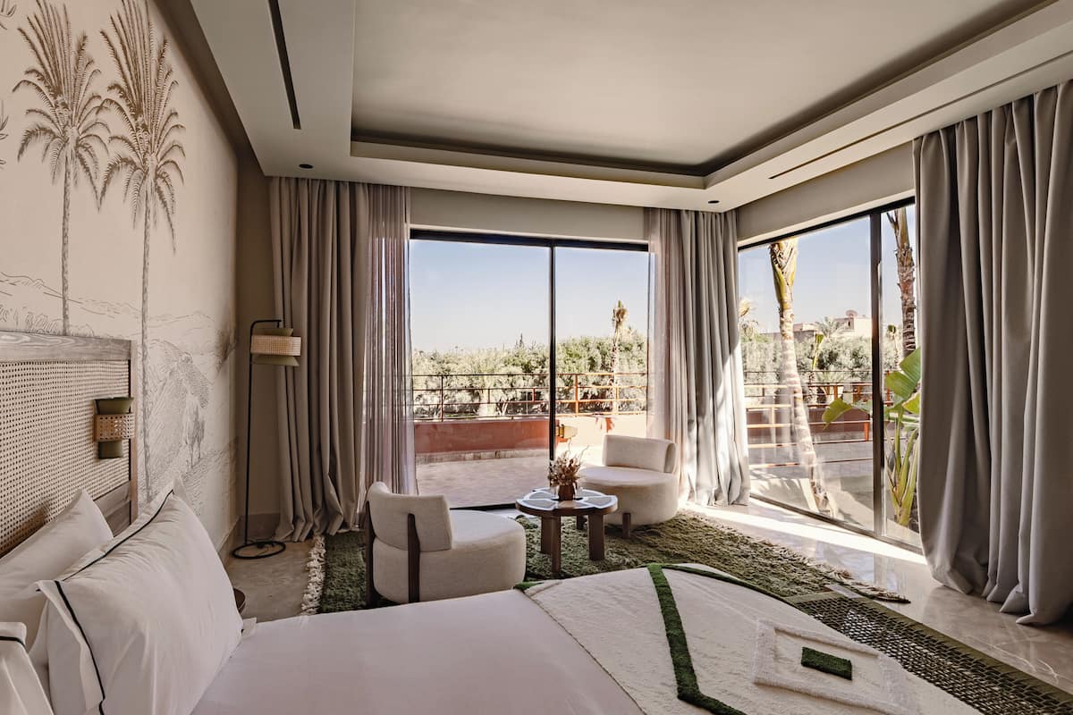 Une chambre d’hôtel avec un lit, deux chaises, une petite table, une lampe et de grandes fenêtres avec des rideaux.