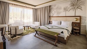 Ein modernes Hotelzimmer mit einem großen Bett, einer Sitzecke mit Stühlen, einem kleinen Tisch und Blick auf Palmen durch das Fenster.
