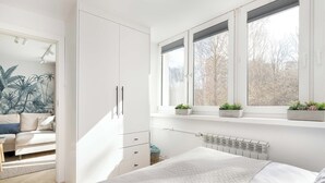 A bedroom with a bed, a radiator, a window with blinds, and a plant on the windowsill. - Cozy Apartment with Balcony | Close to Beach and Station (Gdynia)