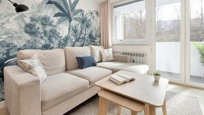A modern living room with a beige sofa, a coffee table, and a large window with a view of trees. - Cozy Apartment with Balcony | Close to Beach and Station (Gdynia)