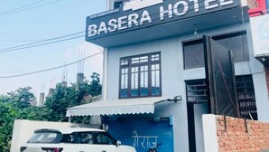 Un immeuble de deux étages sur lequel on peut lire l’inscription « Basera Hotel ». Un VUS blanc est garé devant, et une moto est stationnée sur le côté.