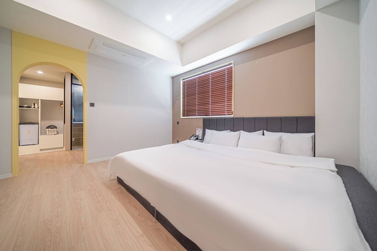 A hotel room with a bed, a window with blinds, a wardrobe, and a bathroom visible through an archway.