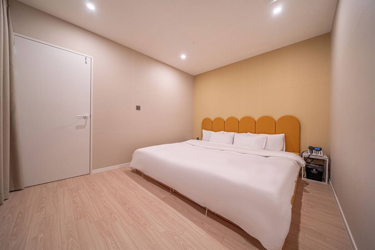 A bedroom with a bed, a bedside table, a nightstand, a door, and a wall-mounted light fixture.