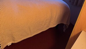 Una habitación de hotel con una cama, almohadas, una mesita de noche, un interruptor de luz en la pared y una ventana con cortinas.