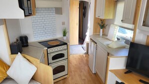 Una kitchenette compacta con estufa, horno y lavabo, un pequeño rincón de descanso con sofá y un televisor sobre una mesa de madera.